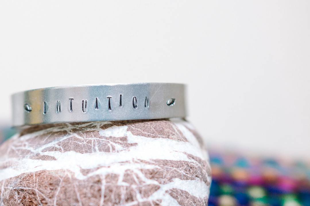 A metal bracelet stamped with the word INUTITION, flanked by two angel wing motifs. It's resting on a pebble and there's a rainbow coloured blanket just visible in the background. Photograph by Carla Watkins Business & Branding Photography at carlawatkins.com